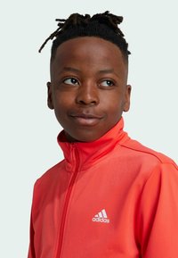 Giacca rossa brillante realizzata in tessuto liscio con colletto alto, dotata di una zip completa e di un logo Adidas bianco sul lato sinistro del petto.