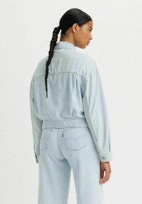 Chaqueta de mezclilla azul clara con una cintura ceñida y mangas largas. La parte trasera presenta costuras verticales. Combinada con pantalones de mezclilla a juego.