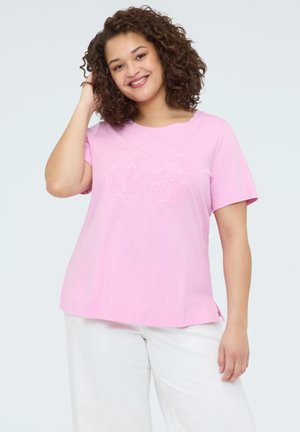 Smilende kvinde med krøllet hår iført en lyserød kortærmet skjorte med tallet 30 og hvide bukser, poserer med hånd nær håret.