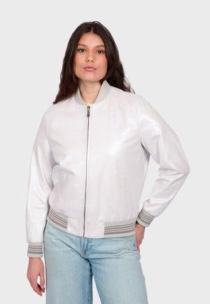 Frau steht mit der Hand in der Hüfte, trägt eine weiße Bomberjacke und hellblaue Jeans vor einem einfarbigen Hintergrund.