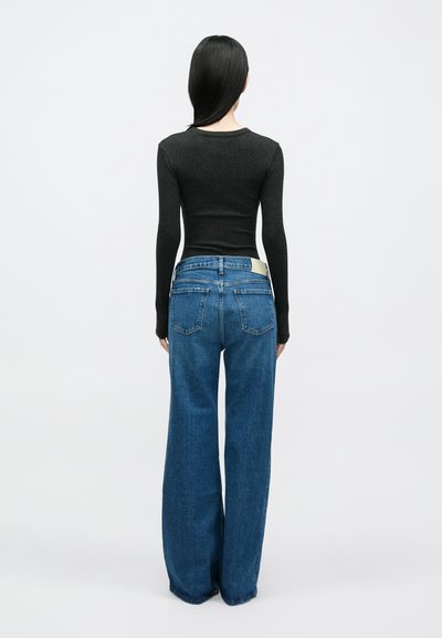 Μαύρο μπλουζάκι με μακριά μανίκια σε συνδυασμό με ψηλό�μεσα, φαρδιά μπλε τζιν παντελόνια. Τα τζιν έχουν τσέπες στο πίσω μέρος και μια ετικέτα ανοιχτού καφέ στη μέση.