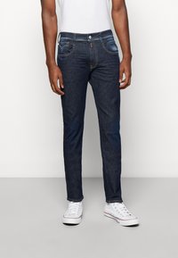 Man som bär mörkblå slim-fit jeans, vit t-shirt och vita sneakers, stående mot en enkel ljus bakgrund.