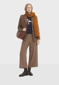 Traje de cuadros marrón con pantalones de pierna ancha, combinado con un suéter azul marino que presenta un gráfico de un pájaro colorido. Bolso de hombro de cuero marrón y bufanda mostaza.