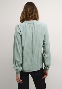 Blusa verde claro con tejido texturizado, que presenta un patrón de cuadrícula, escote fruncido y puños elásticos. Se muestra la vista trasera.