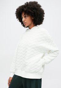 Biała bluza z kapturem o teksturowanym wzorze falistym, z luźnym krojem i ściągaczami na rękawach, w połączeniu z ciemnozielonymi spodniami plisowanymi.