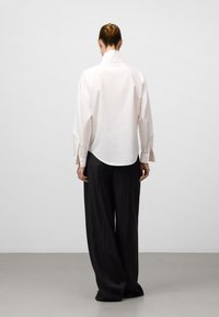 Camicia bianca a maniche lunghe con colletto alto, abbinata a pantaloni neri larghi. La camicia presenta un orlo curvo e polsini con bottoni.