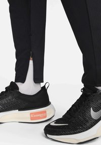 Crne atletske hlače s patentnim zatvaračima na rubovima i teksturiranim završetkom uparene s crnim Nike ZoomX cipelama koje imaju bijeli swoosh i narančastu oznaku.