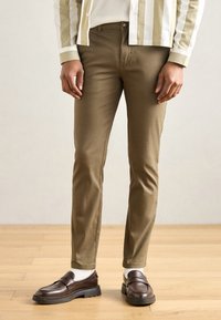 Chino broek van lichtbruin katoenen stof, slim fit, met een knoopsluiting aan de voorkant en zijzakken, gecombineerd met bruine loafers en witte sokken.