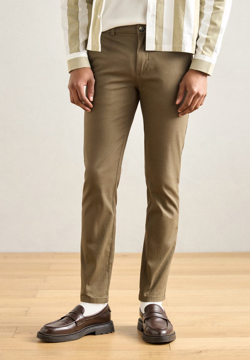 Chino broek van lichtbruin katoenen stof, slim fit, met een knoopsluiting aan de voorkant en zijzakken, gecombineerd met bruine loafers en witte sokken.