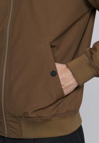 Brown bomber jacket with a zipper, side pocket with a black button, ribbed cuffs, and a smooth, slightly shiny texture.