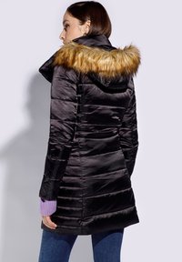 Chaqueta de plumas negra con costuras horizontales, que presenta una capucha de piel sintética marrón claro desmontable y una textura brillante y suave. Mangas moradas visibles.