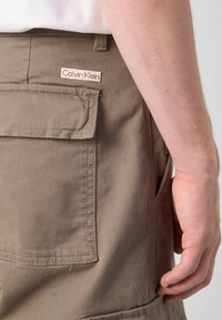 Beige cargoshorts lavet af bomuld med en sidelomme og en baglomme. Har et rødt og hvidt "Calvin Klein" logo-tag.