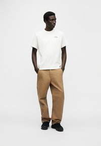 Άσπρο κοντομάνικο μπλουζάκι με μικρό λογότυπο, συνδυασμένο με καφέ φαρδιά παντελόνια και μαύρα αθλητικά παπούτσια. Απλός σχεδιασμός και ουδέτερα χρώματα.