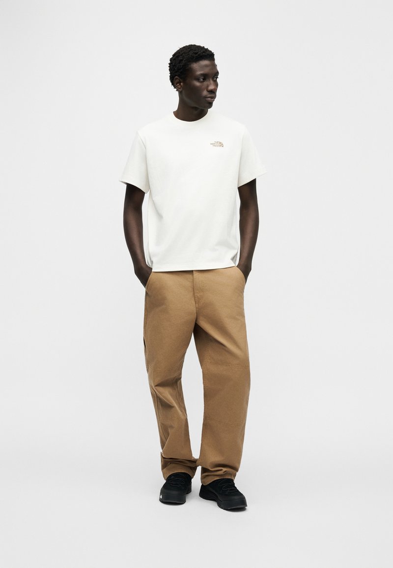 T-shirt blanc à manches courtes avec un petit logo, associé à un pantalon marron ample et des baskets noires. Design simple et couleurs neutres.