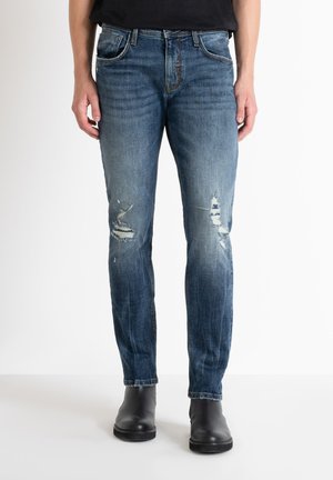 Vaqueros de mezclilla azul deslavado con corte ajustado, que presentan parches desgastados y dobladillos deshilachados. Combinados con botas negras tipo tobillo para crear contraste.