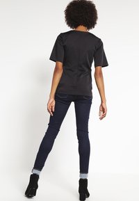 Černý krátký rukávový top s jednoduchým designem, hladkou texturou a zadním zipovým zapínáním, doplněný o tmavě modré upnuté džíny a kotníkové boty.