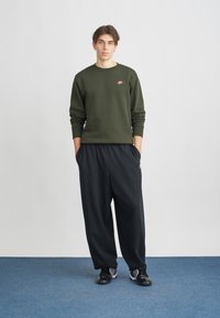 Sweatshirt verde escuro com decote redondo e logotipo da Nike em rosa, combinado com calças largas pretas. A modelo usa sapatos desportivos pretos.