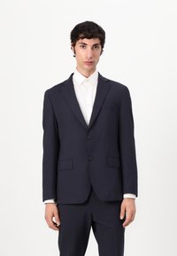 Chaqueta de traje ajustada en azul marino con dos bolsillos frontales, solapas muescas y cierre de dos botones, combinada con una camisa blanca de cuello.