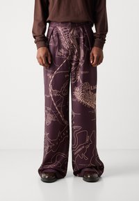 Pantalones de pierna ancha morados con un estampado de mapa en beige. Fabricados en una tela suave con un ajuste relajado y un diseño plisado. Combinándolos con zapatos marrones.