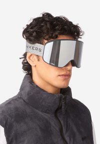 Gogle narciarskie z szerokim, lustrzanym szkłem, szary pasek z tkaniny z napisem "HAWKERS" oraz elegancki, kanciasty design. Noszone z czarną kurtką puchową.