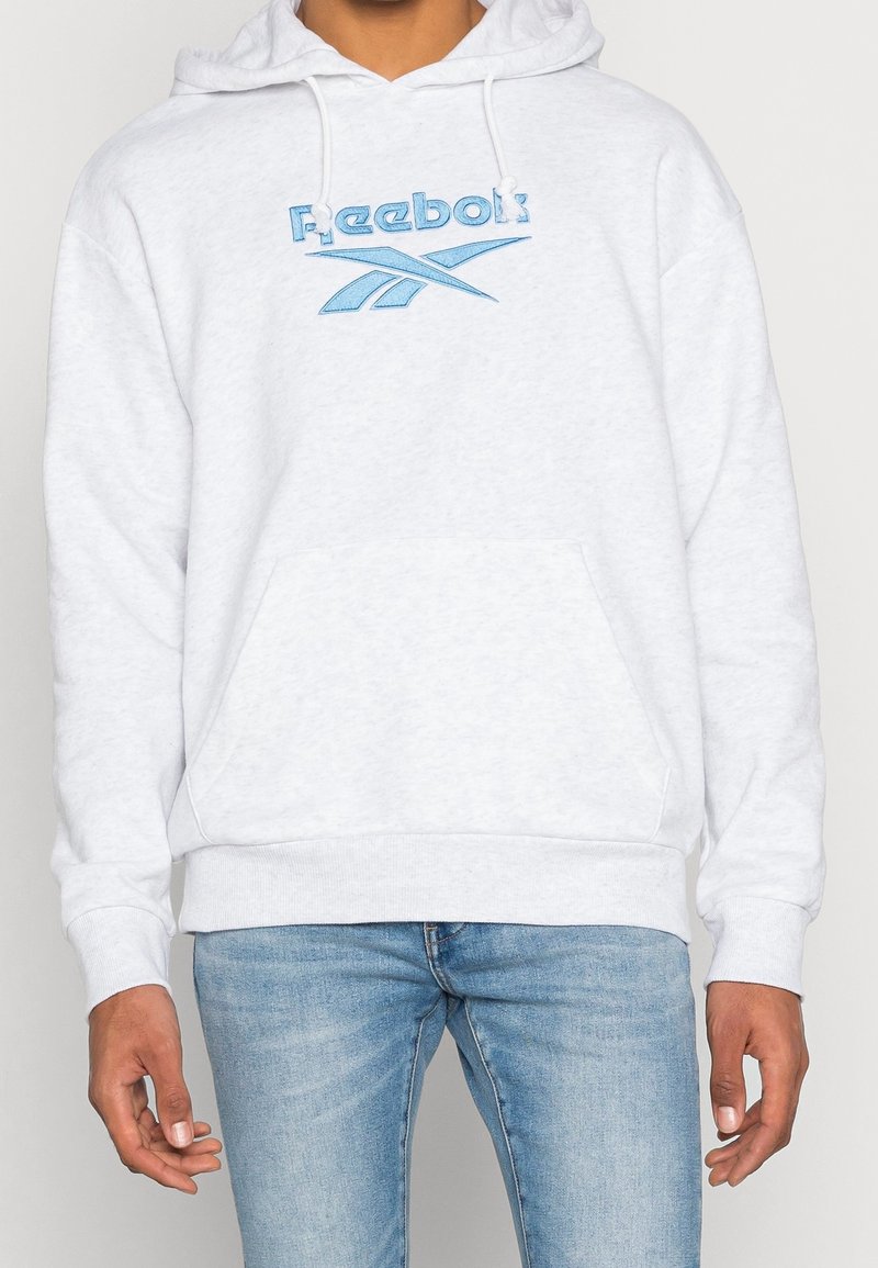 Persona con una sudadera blanca de Reebok con logo azul y vaqueros azul claro, con las manos relajadas a los lados, sobre un fondo liso.