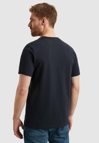 T-paita tummansinisestä puuvillasta, jossa on pyöreä kaula-aukko, lyhyet hihat ja pelkkä takasuunnittelu, yhdistettynä vaaleansinihin farkkuihin.