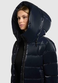 Giubbotto imbottito blu navy con finitura lucida, cappuccio oversize, chiusura con zip e texture trapuntata. Presenta lacci per una vestibilità regolabile.