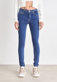 Jeans ajustados azul con una cintura de media altura, bolsillos frontales y un ligero estiramiento. Combinados con zapatillas deportivas beige. Textura suave.