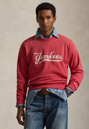 Mann kledd i rød "Yankees" genser over dongeri skjorte og jeans, stående med en hånd i lommen foran ensfarget bakgrunn.