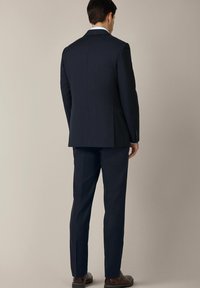 Granatowa marynarka o gładkiej fakturze, z pojedynczym rozcięciem i dwiema guzikami, zestawiona z dopasowanymi spodniami i brązowymi butami, widok z tyłu.