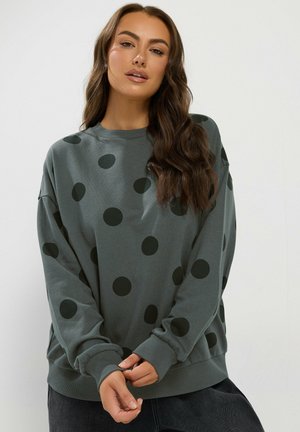 Femme aux longs cheveux ondulés portant un sweat-shirt gris avec de grands pois noirs, debout devant un arrière-plan blanc uni.