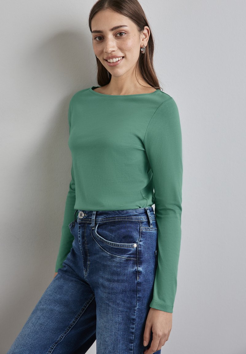 Langärmliges, grünes Baumwolloberteil kombiniert mit blauen, hochgeschnittenen Jeans, die einen klassischen Schnitt und seitliche Taschen aufweisen.