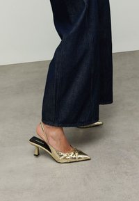 Escarpins slingback métalliques dorés avec bout pointu, ornés de nœuds décoratifs et d'un petit talon bloc. Portés avec un denim large foncé.