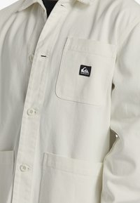 Light cream canvas jacket with a front pocket, button closure, and a small black logo patch. Features a collared neck and a textured finish.