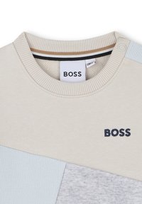 Sweatshirt beige clair avec un col côtelé, présentant des accents de panneaux bleu et gris. Comprend un logo "BOSS" en couture navy.