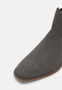 Gros plan de l'avant et du côté d'une bottine en daim gris foncé avec une semelle marron sur fond blanc.