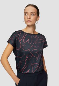 Kurze ärmellose marineblaue Bluse mit einem roten Herzmuster, glatter Textur und weitem Ausschnitt, kombiniert mit dunklen Hosen.