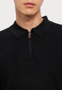 Polo shirt nero a coste con colletto zip e dettagli in metallo argentato. La texture del tessuto è prominente, creando un aspetto strutturato.