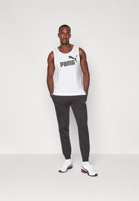 Biela tielko bez rukávov s čiernym logom "PUMA", skombinované s tmavošedými teplákmi a bielymi športovými topánkami s červenými a čiernymi akcentmi.