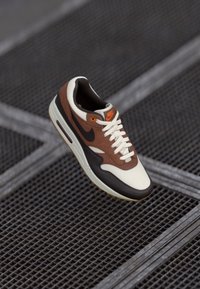 Sapatilha Nike branca, castanha e preta com logotipo laranja, atacadores atados, posicionada acima de uma superfície de rede metálica escura.