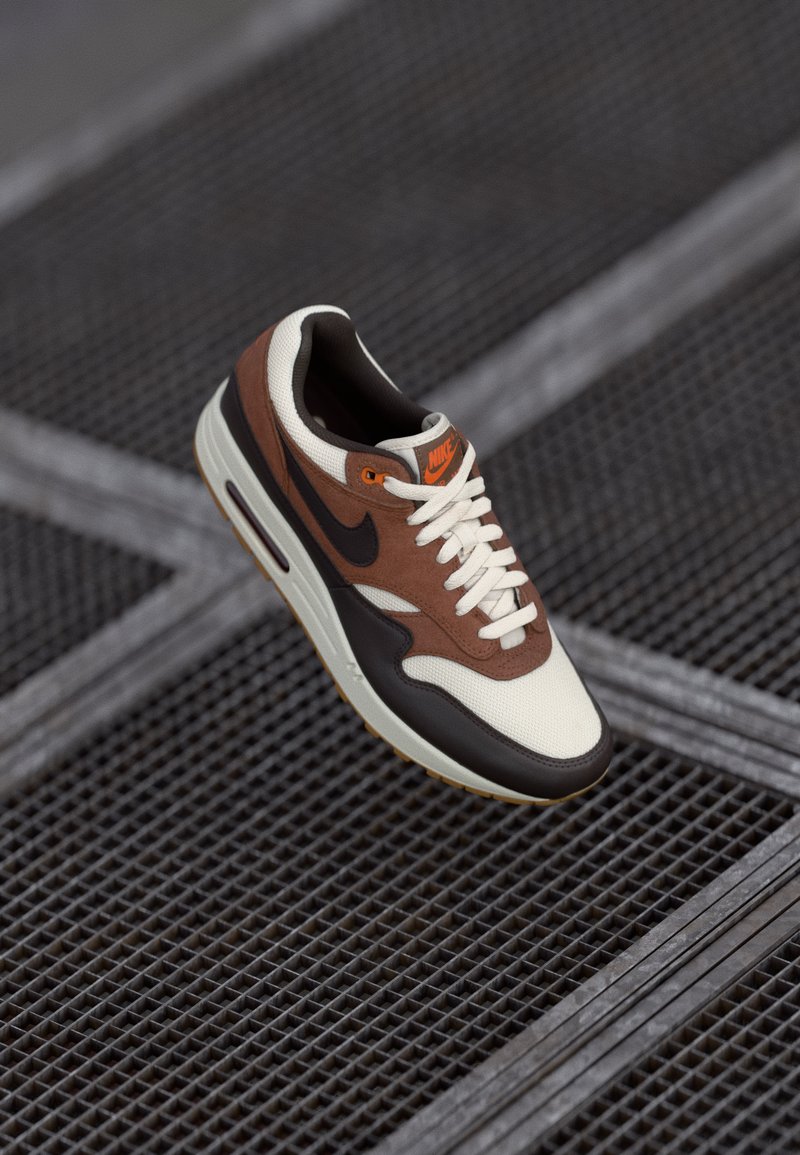 Sapatilha Nike branca, castanha e preta com logotipo laranja, atacadores atados, posicionada acima de uma superfície de rede metálica escura.
