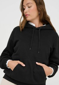 Czarna bluza z kapturem z kieszenią z przodu, regulowanymi sznurkami i ściśniętymi mankietami. Materiał wydaje się miękki, z luźnym krojem i minimalistycznym designem.