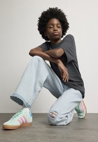 Ungdom med afrofrisyre som står på kne, iført en m�ørk t-skjorte, lyse ribbede jeans, hvite sokker, og mintgrønne sneakers med rosa striper.
