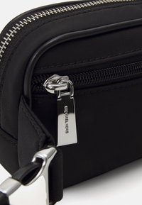 Musta nylon crossbody -laukkua, jossa on hopeista metalliosaa ja vetoketjullinen sulku. Varustettu tuotemerkillä ja teksturoidulla pintakuvioinnilla.