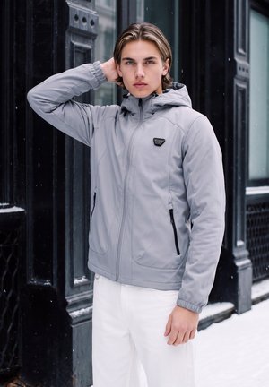 Jeune homme en veste à capuche grise et pantalon blanc se tient dans une rue enneigée, une main touchant ses cheveux près d'une façade noire de bâtiment.