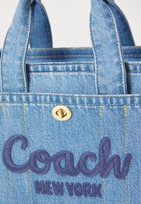 Farmeraruház táska kék szövetből, hímzett "Coach New York" logóval sötétkék színben, aranyszínű fém kiegészítőkkel és sárga varrási díszítésekkel.