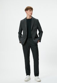 Männlicher Model trägt einen grauen strukturierten Blazer, ein schwarzes Rundhalsshirt und schwarze Hose. Die Schuhe sind weiße Sneaker. Der Model steht mit den Händen in den Taschen.