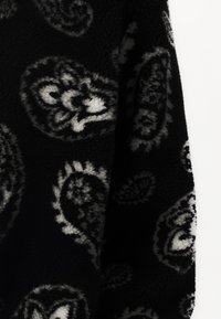 Svart fleece-stoff med et hvitt paisley-mønster. Teksturen er myk og fluffy, med varierende nyanser av grått i designdetaljene.
