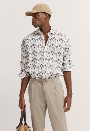 Short-sleeved shirt in light fabric featuring a colorful bird pattern, paired with beige trousers and a tan cap. Holding a bag.