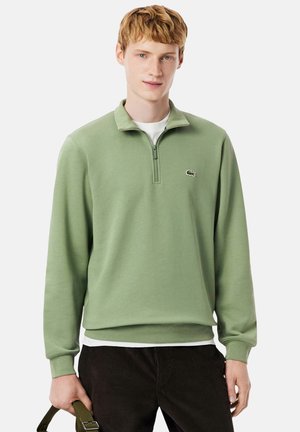 Joven con sudadera de cremallera corta verde claro, camiseta blanca debajo y pantalones oscuros, sosteniendo una correa verde con la mano izquierda.